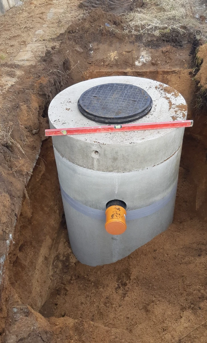 Szambo betonowe w trakcie montażu w wykopie, widoczny betonowy zbiornik z pokrywą i rurą odpływową