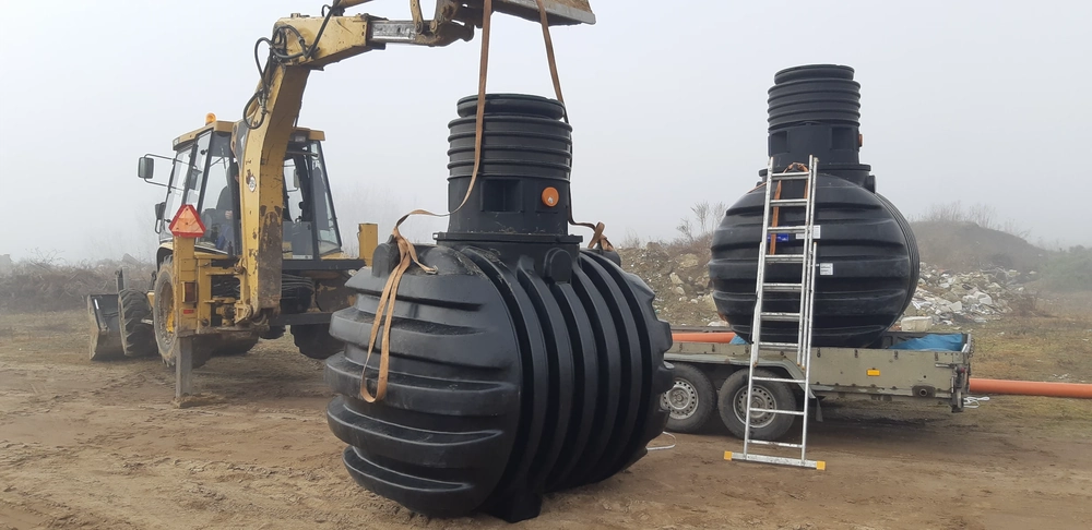 Ładowarka podnosi czarny plastikowy zbiornik podziemny na placu budowy, obok widać drabinę i drugi zbiornik