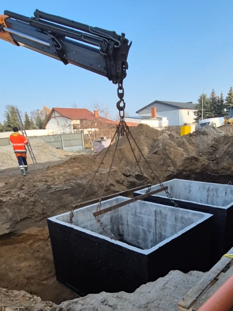 Betonowe sekcje szamba betonowego opuszczane dźwigiem do wykopu na placu budowy, widoczny robotnik w pomarańczowej kamizelce