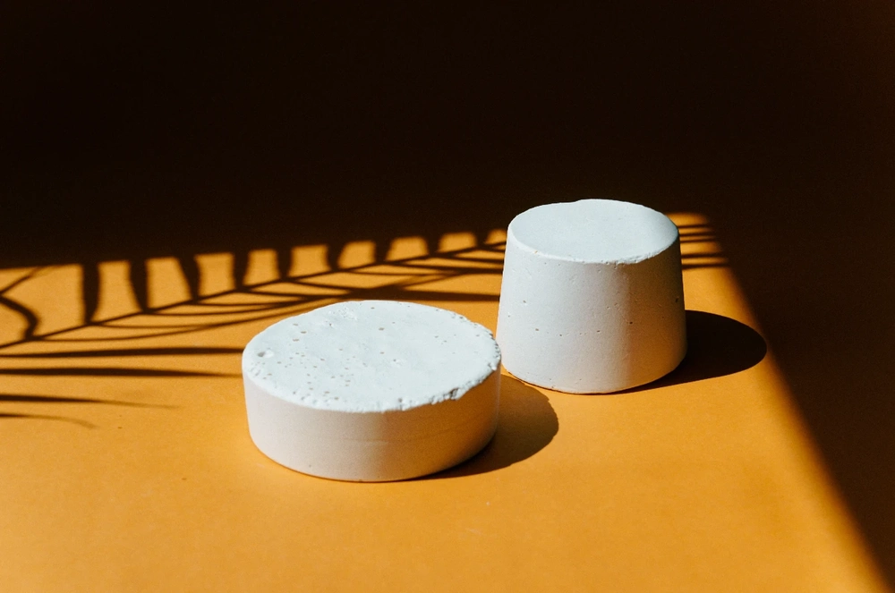 Minimalistyczne ułożenie dwóch białych walcowatych bloczków betonowych na pomarańczowej powierzchni z cieniem liści