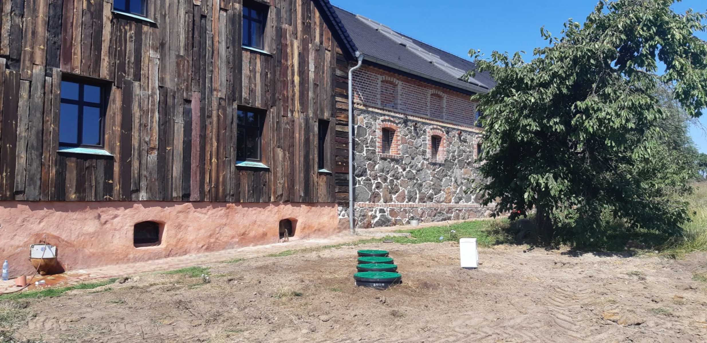 Instalacja przydomowej oczyszczalni ścieków z zielonymi włazami na tle drewniano-kamiennego budynku