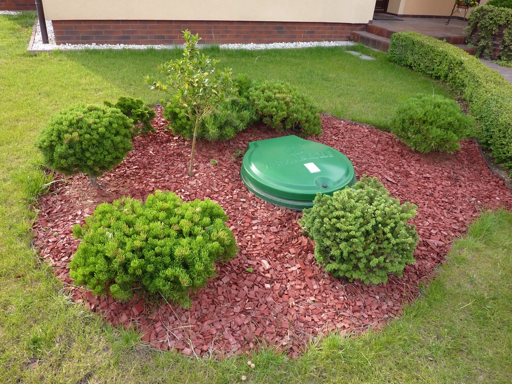 Zielony plastikowy właz przydomowej oczyszczalni ścieków osadzony w czerwonej korze, wokół zadbane iglaki i trawnik